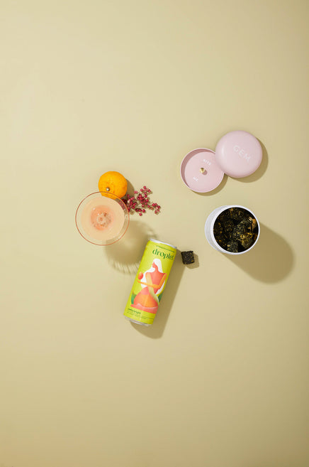 a can of pretty bright lying flat beside a cocktail and a candle artistically presented