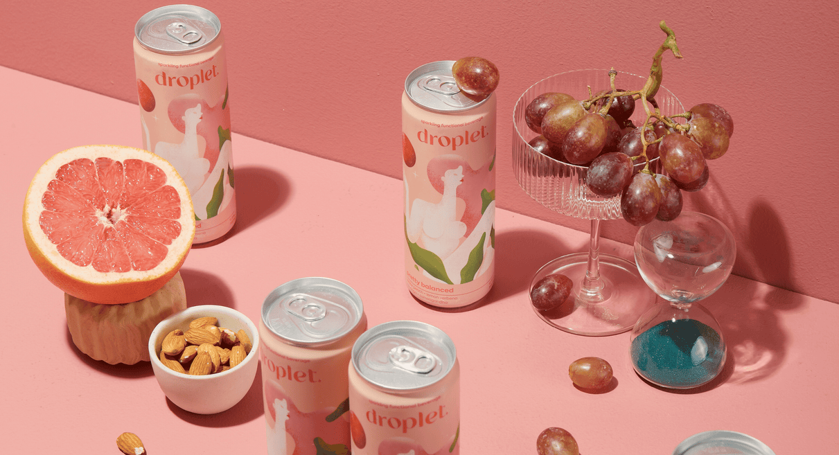 cans of pretty balanced artistically laid out with grapes, wine glasses, grapefruit and almonds to create a luxurious scene against a light pink backdrop