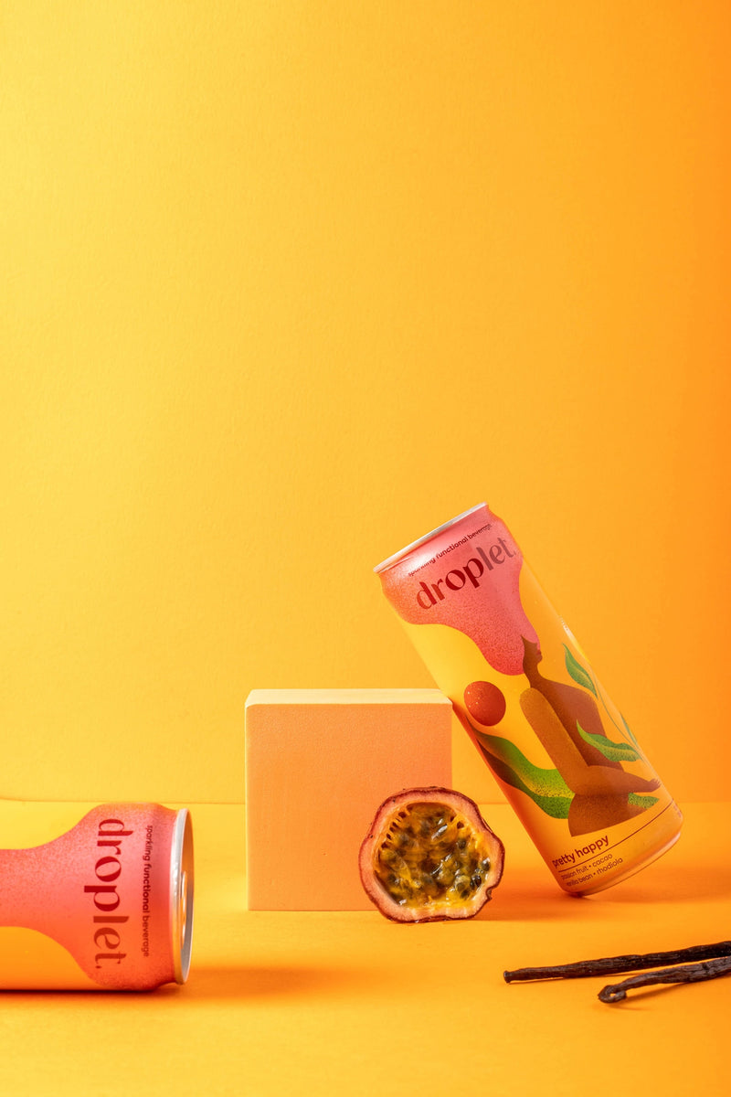 two cans of pretty happy against a rich yellow backdrop and passionfruit and vanilla bean in the shot